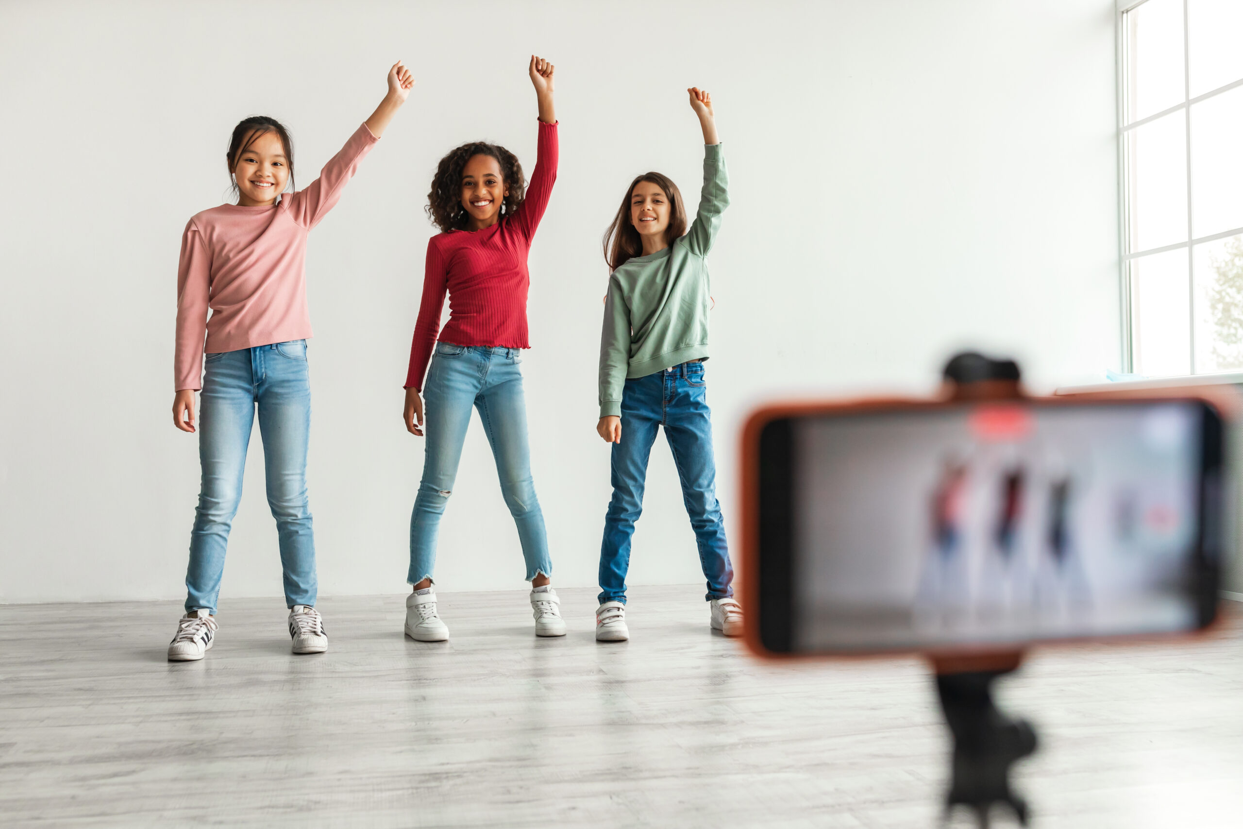 Three Diverse Blogger Girls Making Video For Blog On Smartphone Raising Arms Smiling To Camera Indoors. Kids Posing In Front Of Mobile Phone. Selective Focus